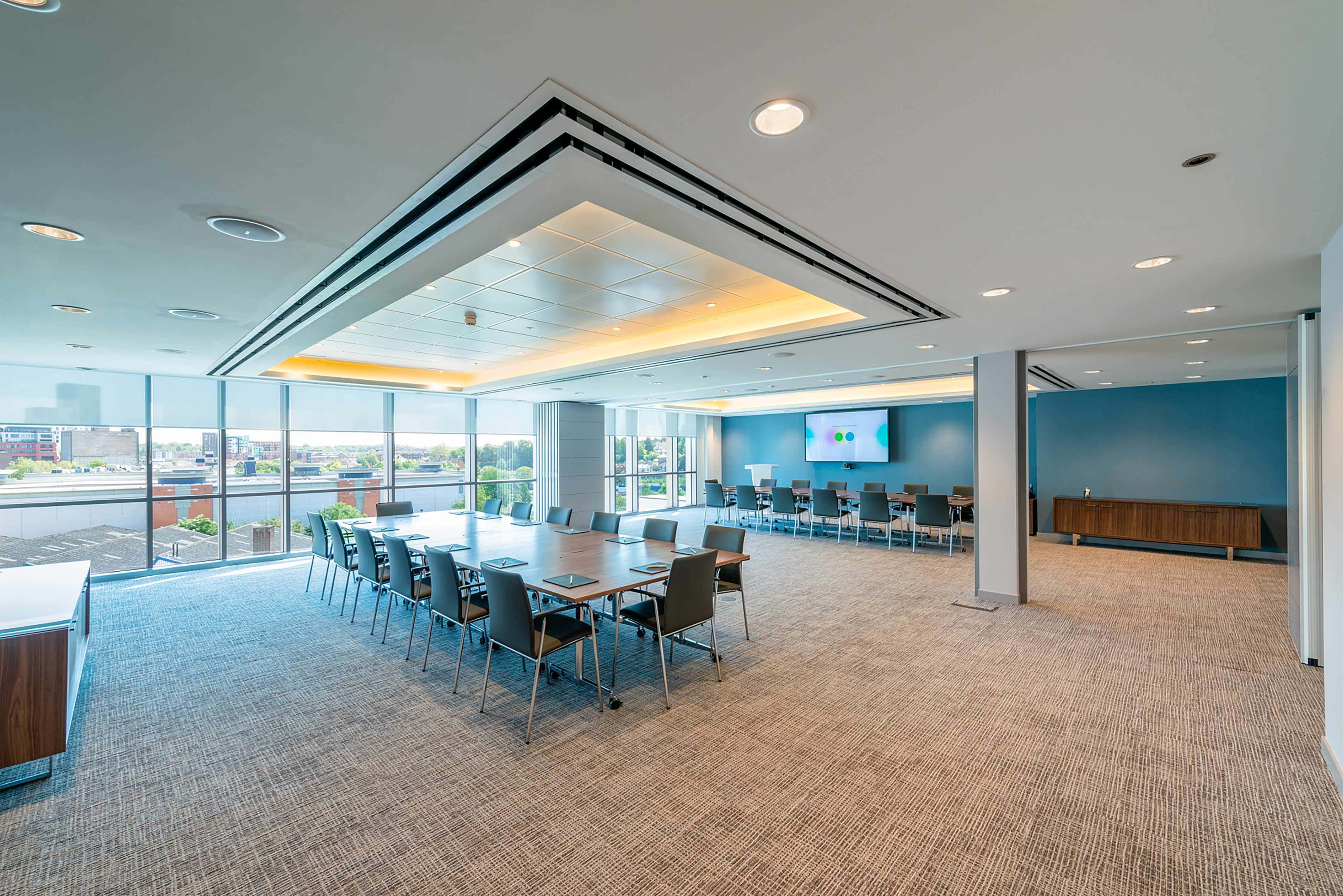 Conference room with large rectangular table, surrounded by chairs, facing a wall-mounted screen displaying two colored circles, in a bright, modern space with a city view through large windows.
