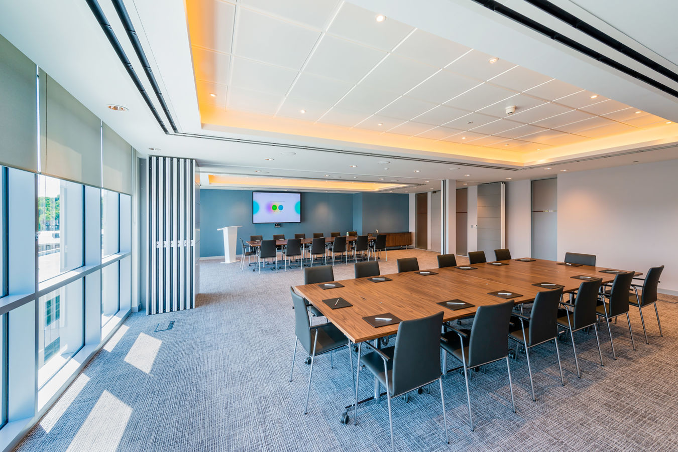modern meeting room with a large wooden table, black chairs, a presentation screen, and large windows