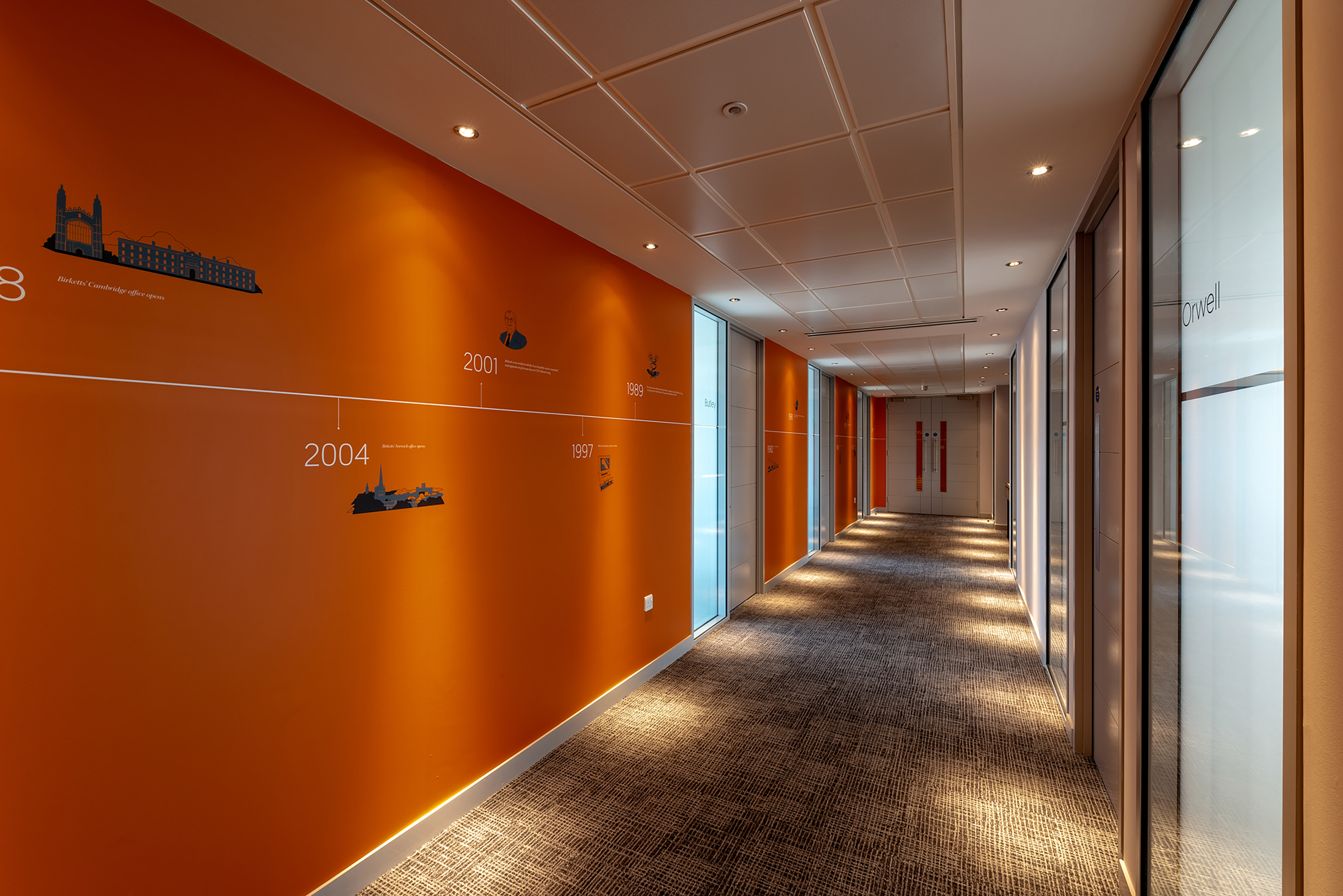 Orange hallway features a timeline with years and illustrated events on the left wall, while glass doors on the right display the name "Orwell," in a modern office setting.