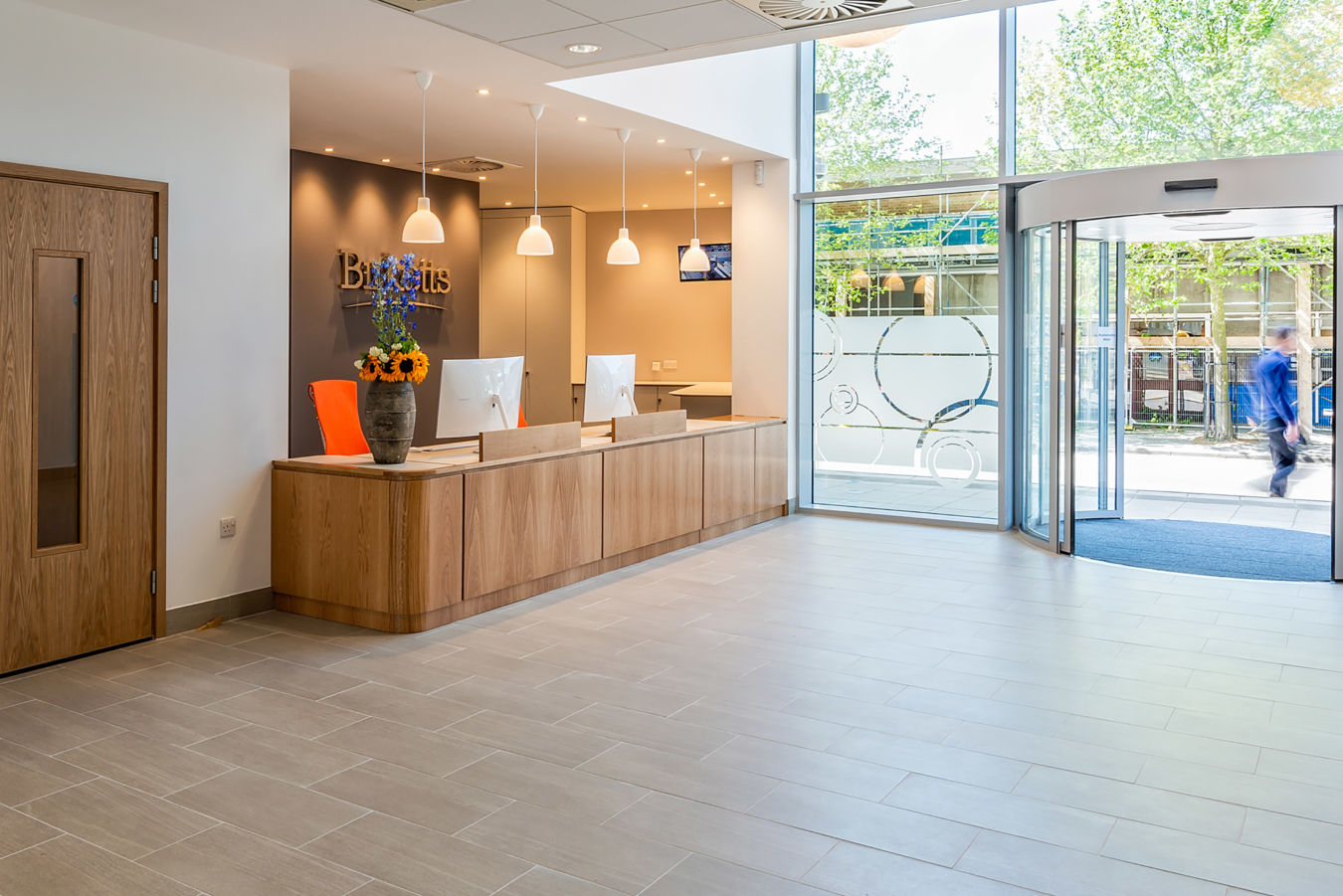 Reception desk with computers and a flower vase; "Birketts" text on wall; glass entrance reveals outdoor street scene; bright, modern office environment with pendant lights and wooden door.