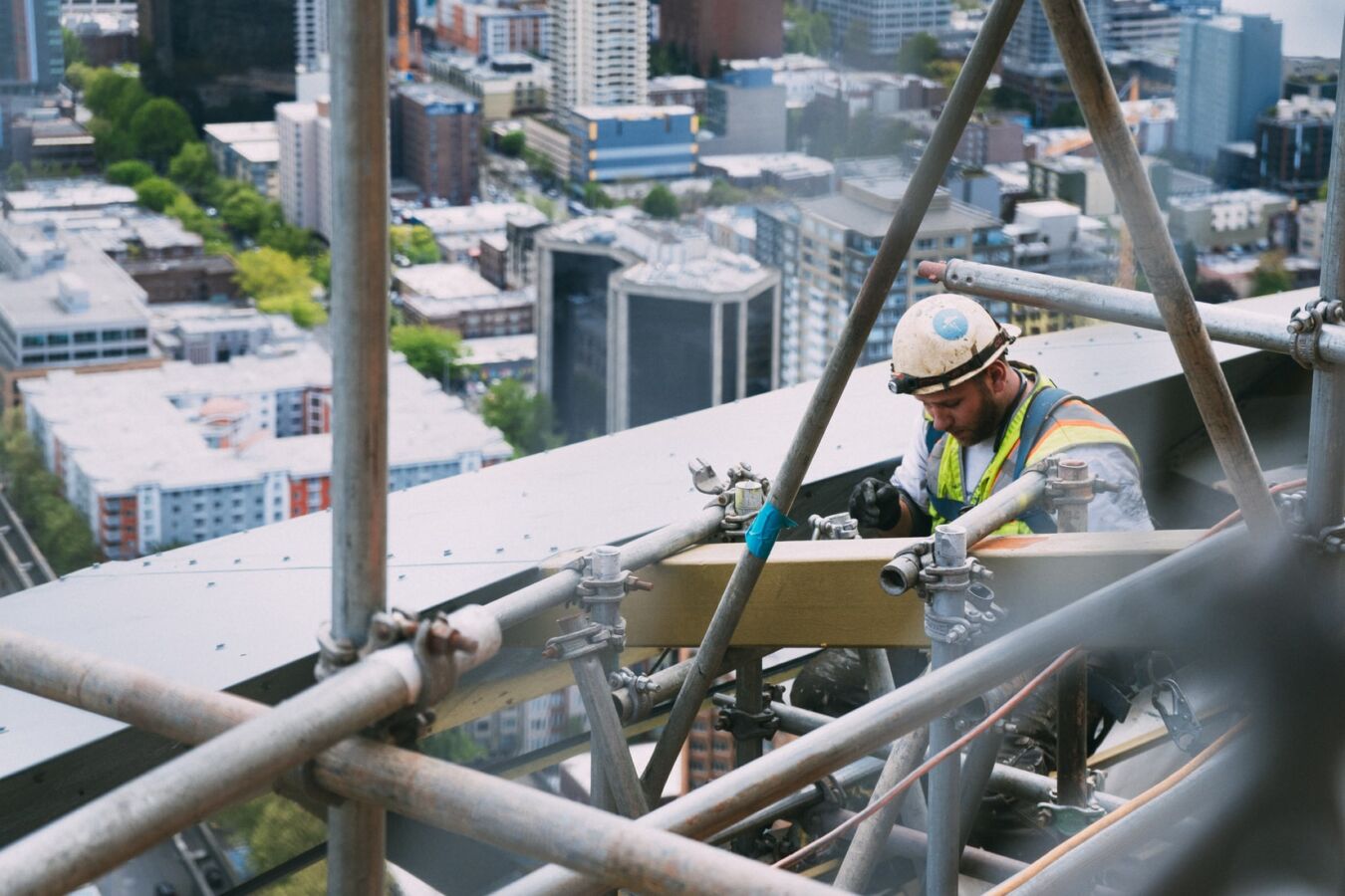 Construction worker repairs scaffolding on a skyscraper, using tools while wearing a hard hat and safety vest; urban cityscape visible below.