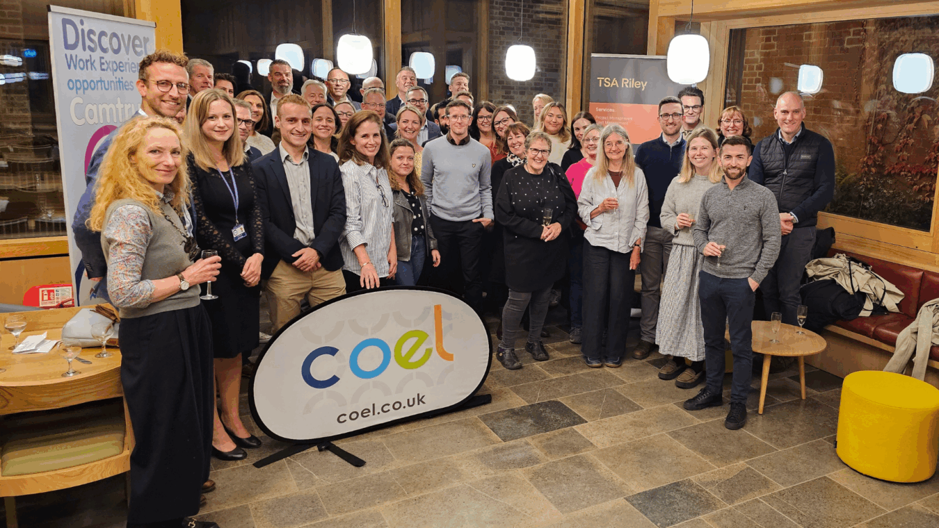 Group of people smiling and posing indoors next to banners reading "Discover Work Experience opportunities Camtruf" and "TSA Riley." A sign with "coel.co.uk" is in front, suggesting a formal gathering.