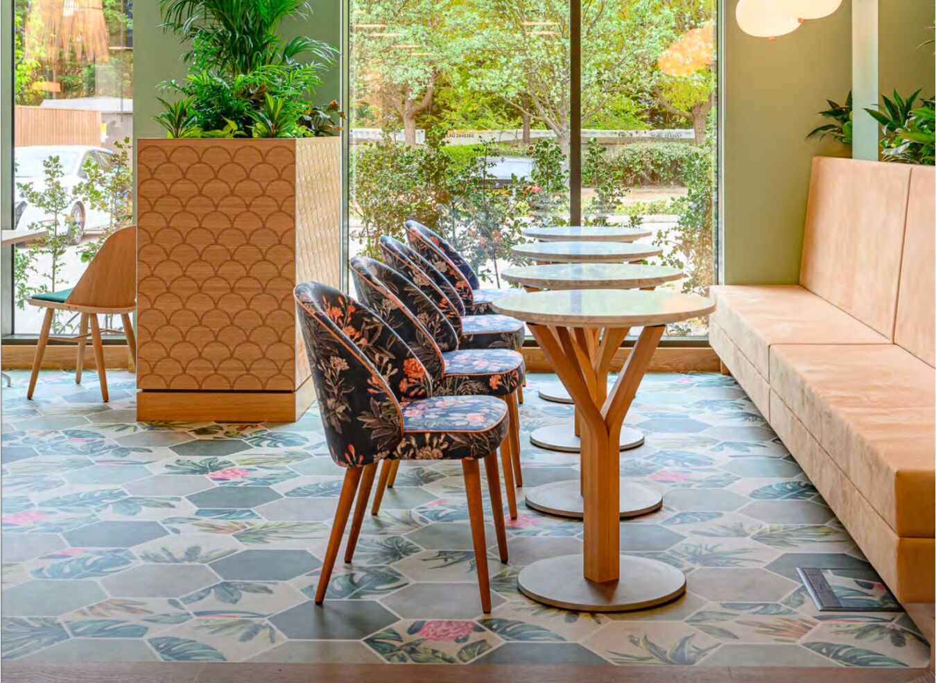 Floral upholstered chairs line wooden tables on a patterned floor in a bright cafe, set against large windows with views of greenery and a wood-paneled planter.