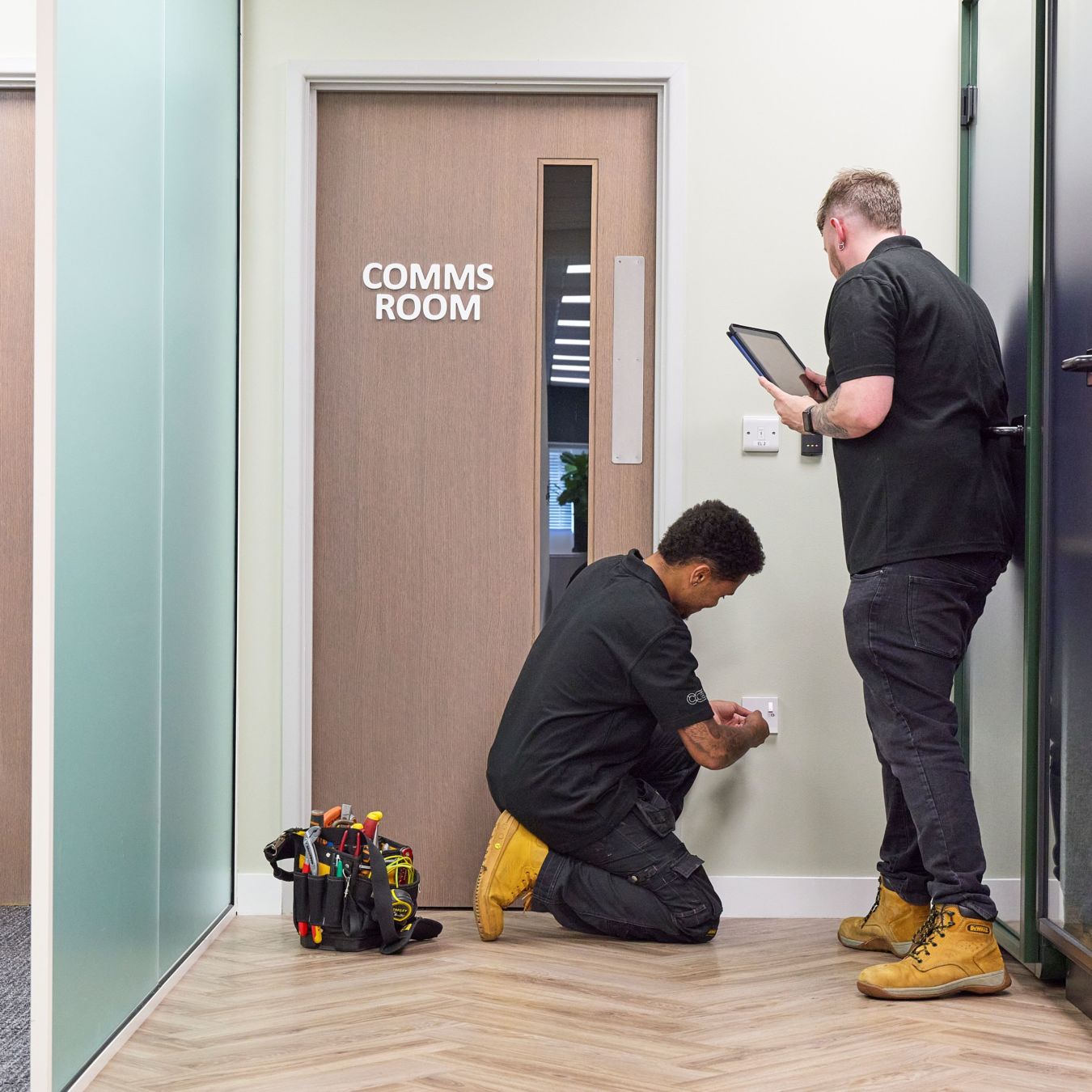 Two technicians work near a wall socket; one kneels adjusting it, the other holds a tablet. A tool bag is on the floor beside them, next to a "COMMS ROOM" door.