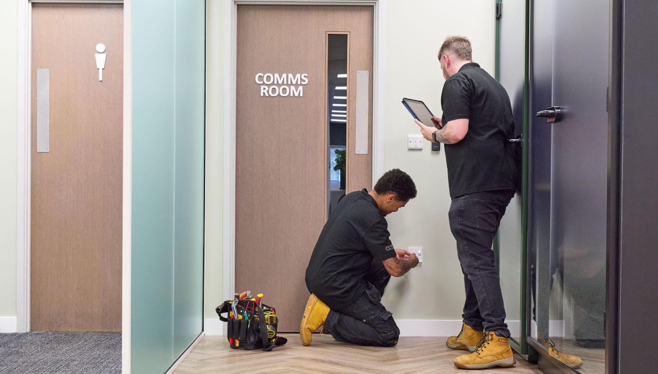 Two technicians work near a wall socket; one kneels adjusting it, the other holds a tablet. A tool bag is on the floor beside them, next to a "COMMS ROOM" door.