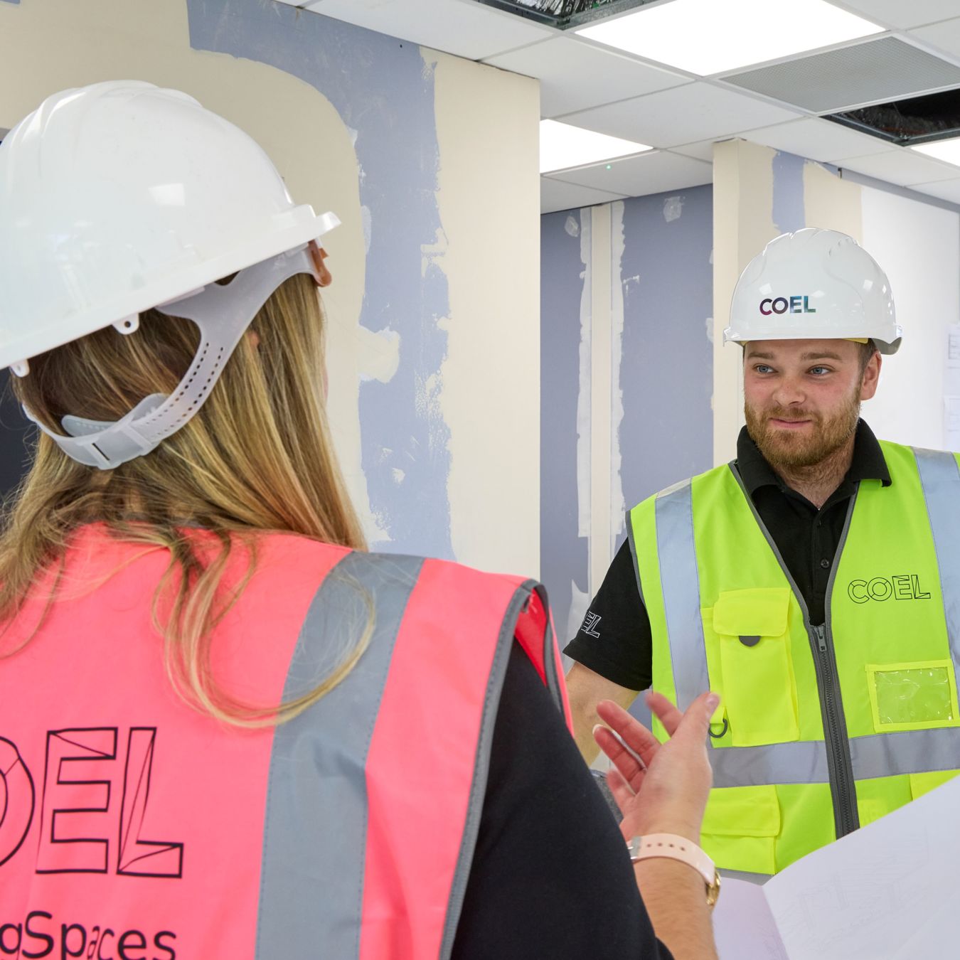 Two construction workers wearing hard hats and high-visibility vests converse inside a partially finished building. "COEL" is visible on their vests. The walls are partially painted.