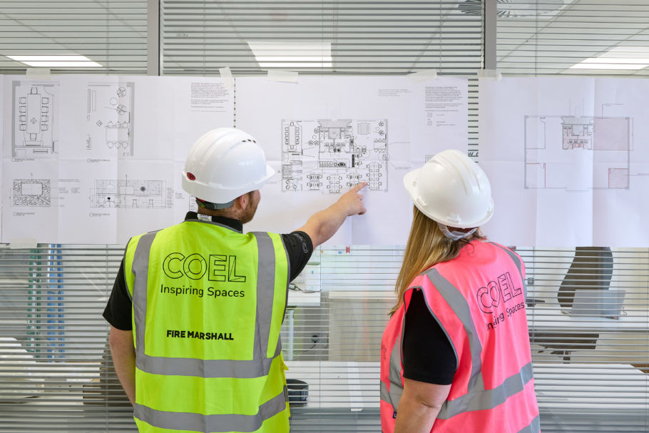 Two people wearing safety vests and helmets discuss architectural plans taped to a glass wall. The atmosphere is focused and collaborative