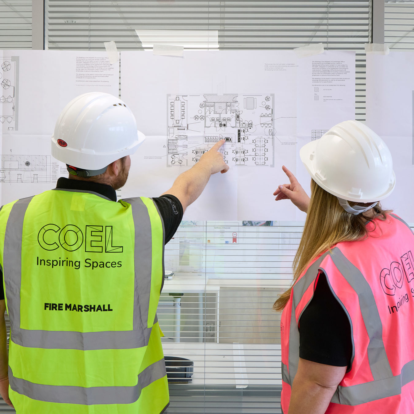 Two construction workers, wearing helmets and vests labeled "COEL Inspiring Spaces," discuss blueprints on a glass wall in an office setting.