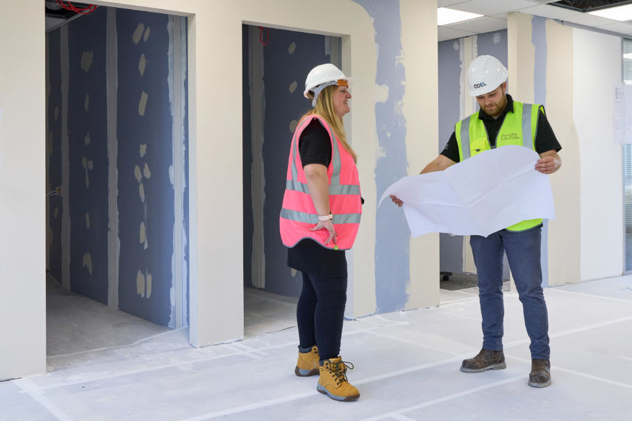 Two construction workers, one in a pink vest, one in a green vest, with hard hats, discuss blueprints in a partially completed interior space