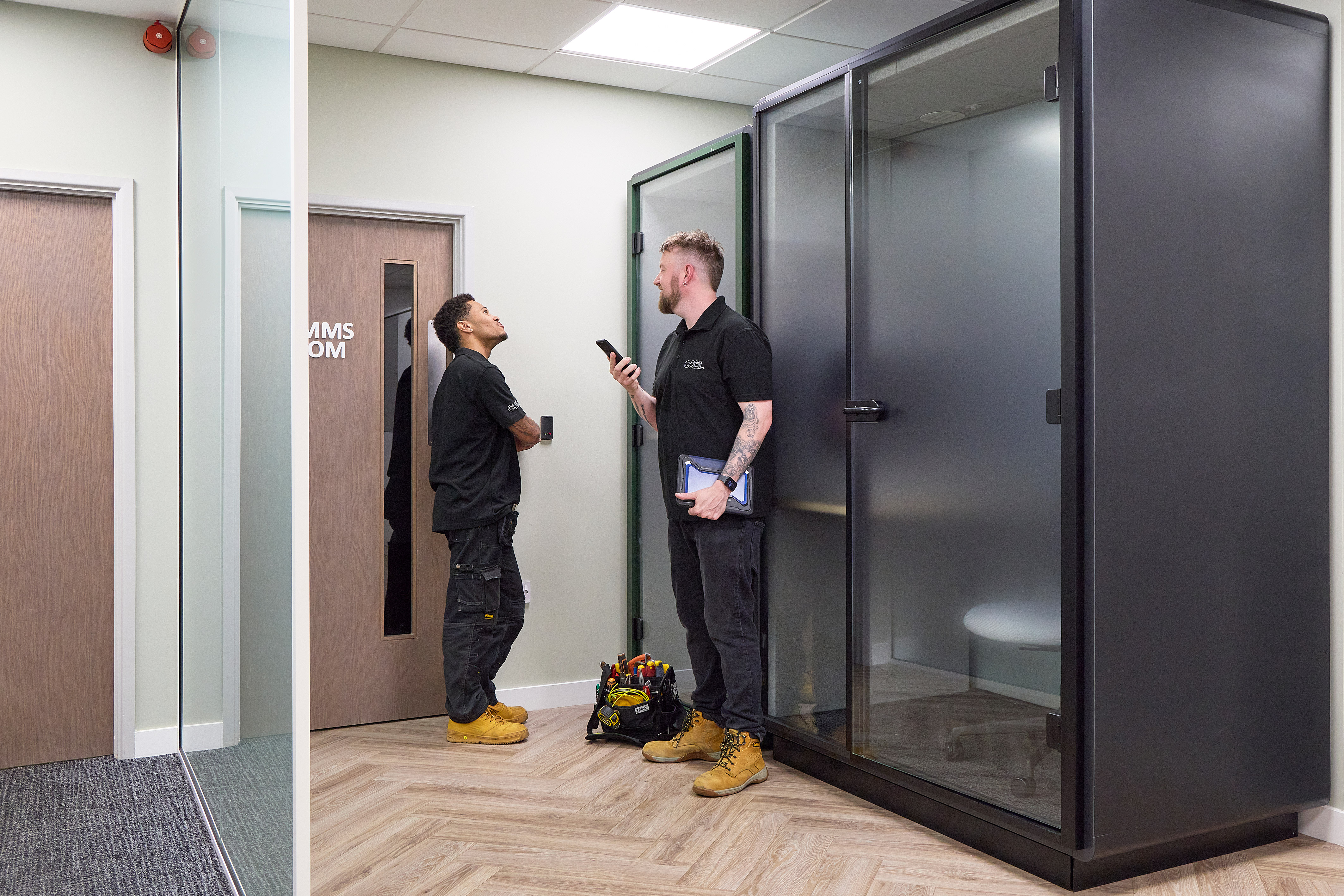 Two workers in black shirts converse in an office hallway. One holds a phone, the other leans against a door labeled "COMMS ROOM." A tool bag sits on the floor.