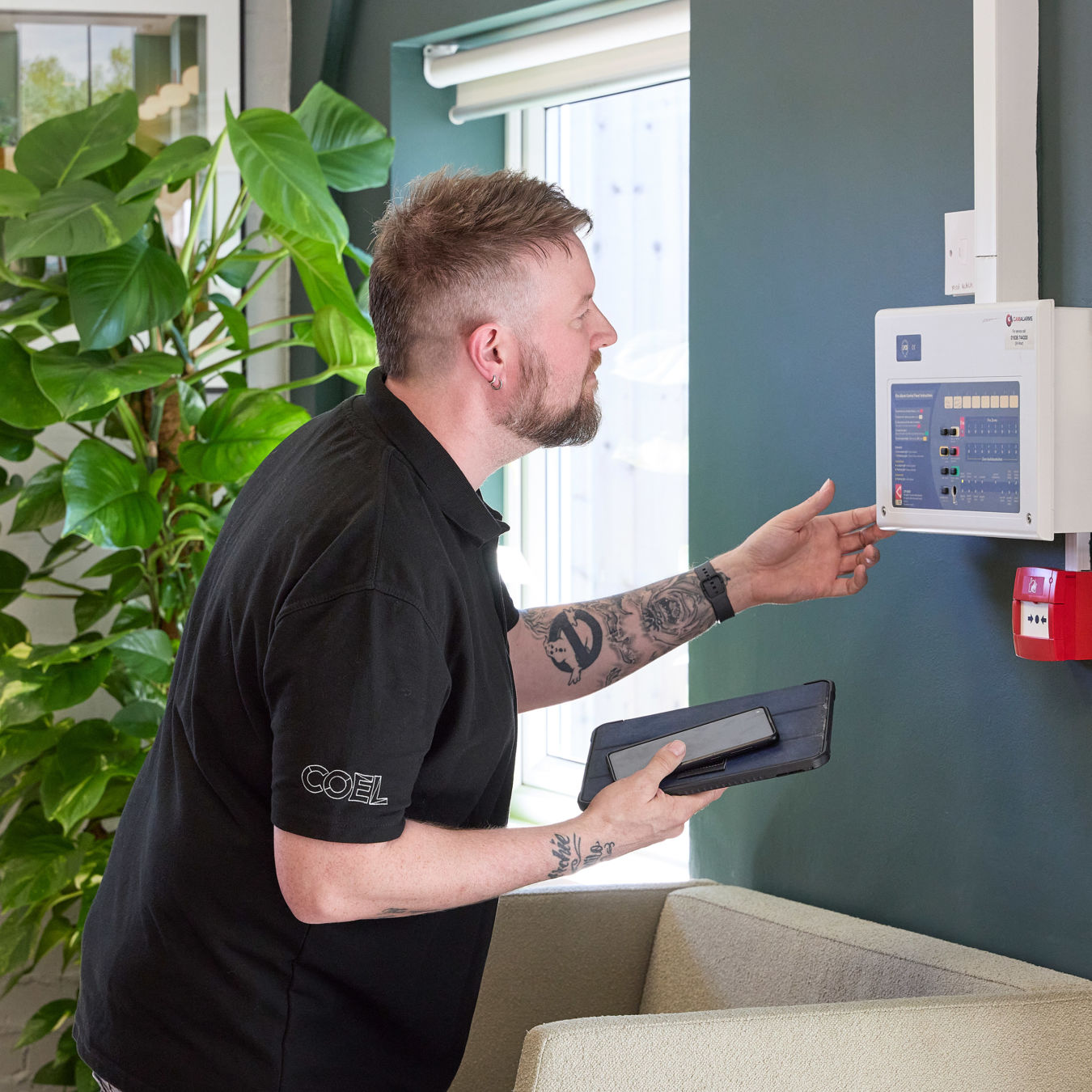 A man examines a control panel mounted on a green wall, holding a tablet, with a large plant nearby and sunlight streaming through a window. The shirt text reads "COEL."