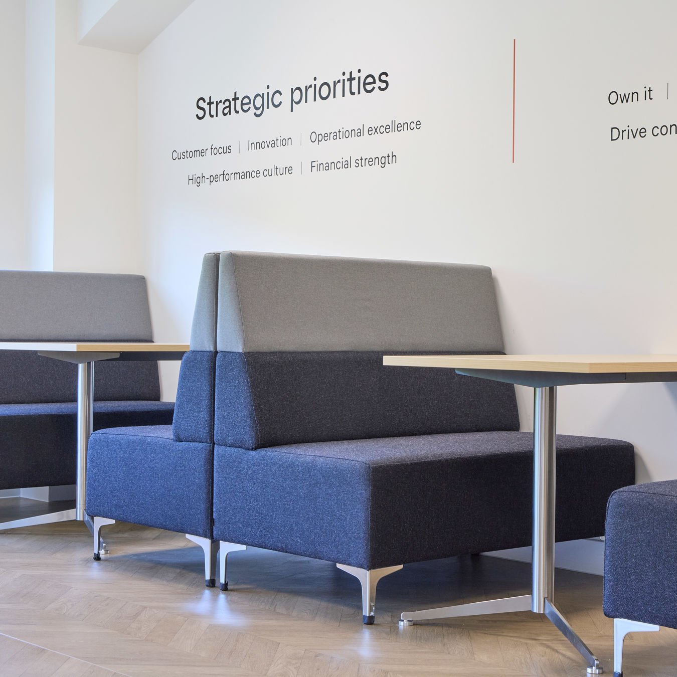 Modern office seating area with gray cushioned booths and wooden tables against a white wall displaying "Strategic priorities". Bright and inviting.