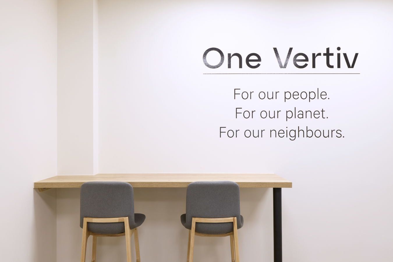 minimalist office nook with a wooden desk, two grey chairs, and a "One Vertiv" mission statement on the white wall