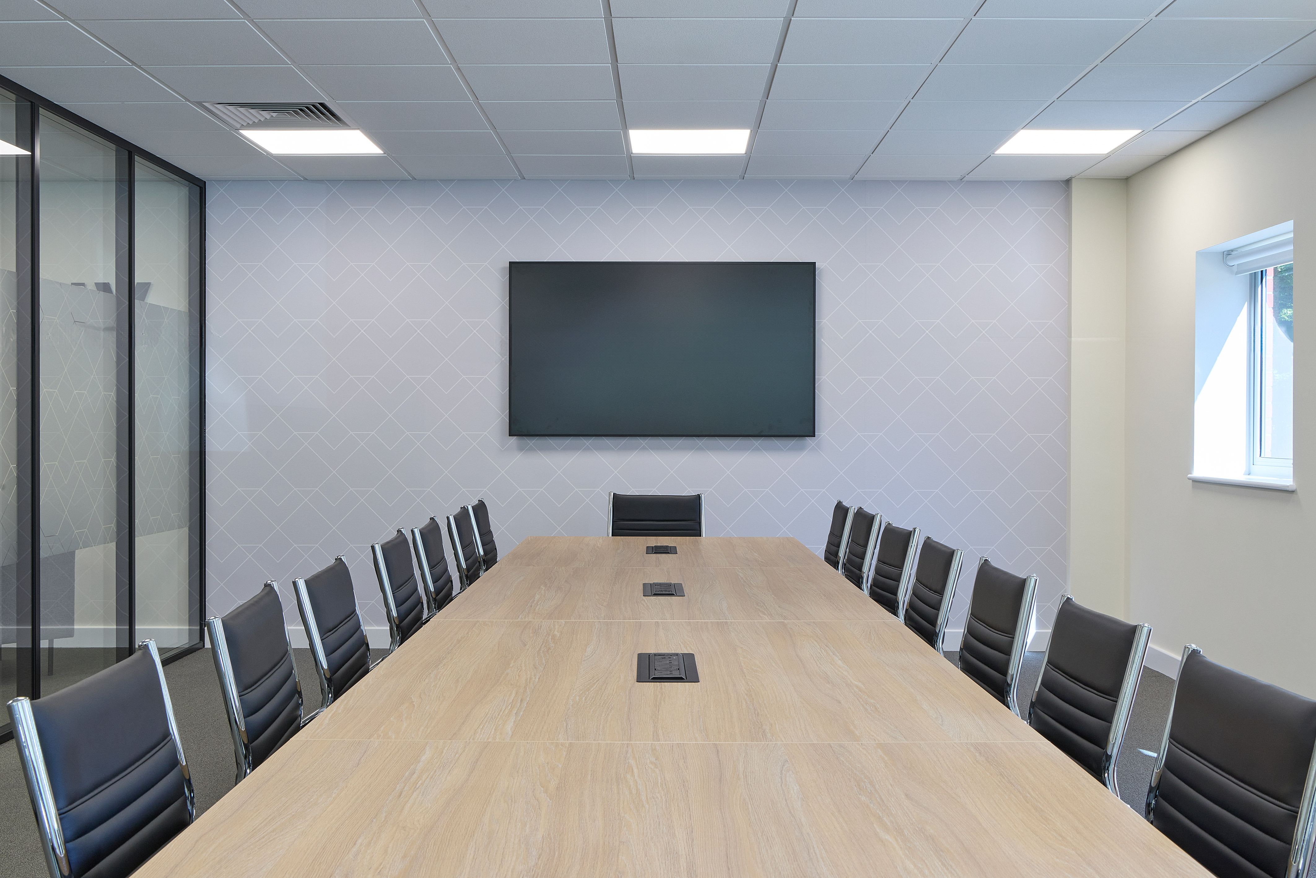 Modern conference room with a large wooden table surrounded by black leather chairs. A blank screen on the wall and a window offering natural light