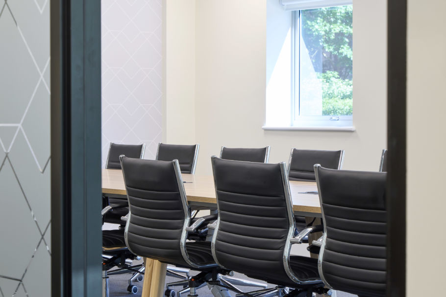 Modern conference room with eight black leather chairs around a wooden table. A window with a view of green foliage adds natural light. Calm ambiance