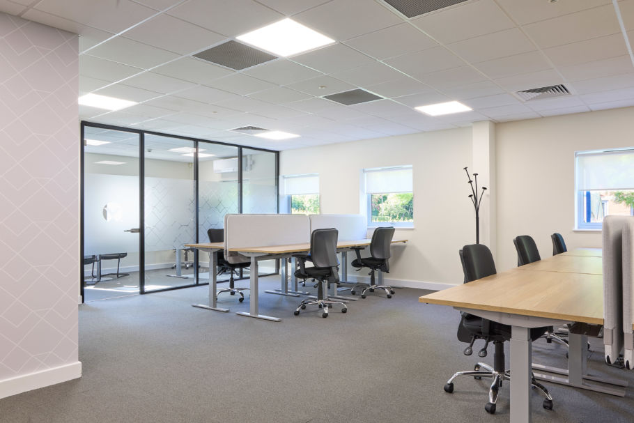 Modern office space with desks and chairs arranged in rows, a glass-walled meeting room, and light streaming through windows. The atmosphere is clean and minimalistic