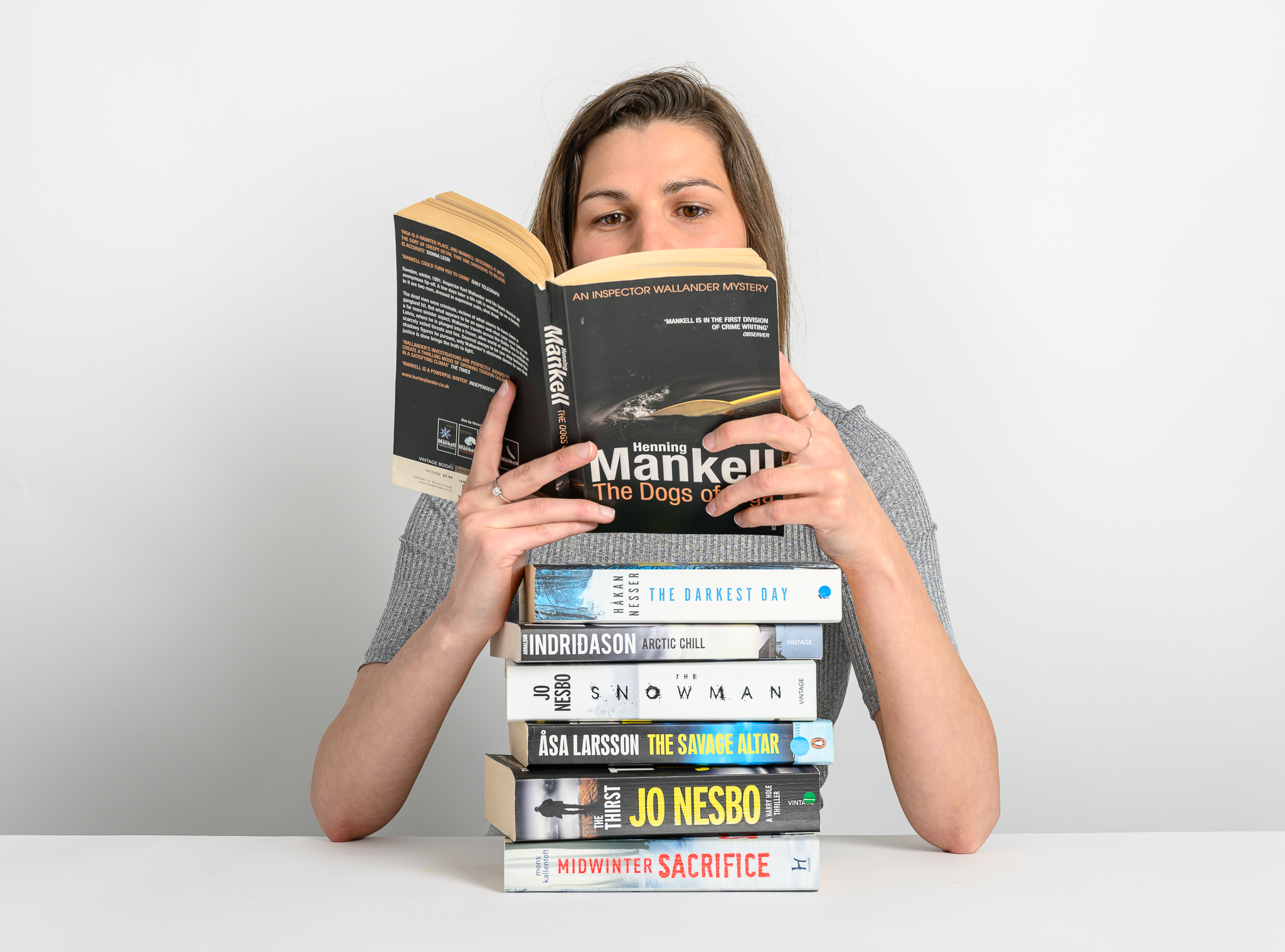 A person reads "Henning Mankell: The Dogs of Riga" over a stack of crime novels, titled "The Darkest Day," "Arctic Chill," "The Snowman," "The Savage Altar," "The Thirst," "Midwinter Sacrifice," on a white table.
