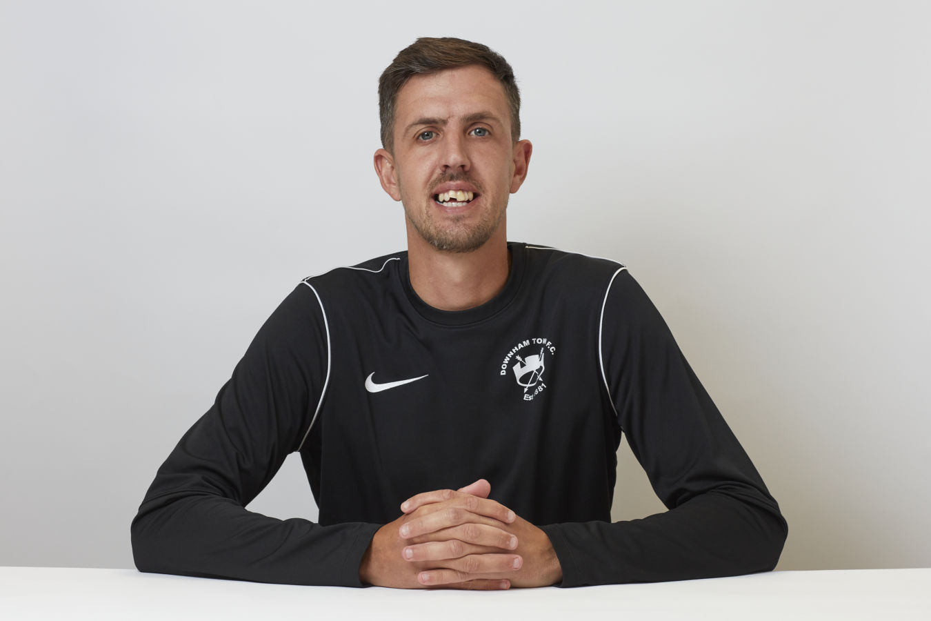 A person sits at a table, wearing a black Nike sports shirt with "DOWNHAM TOWN FC EST 1881" logo, hands clasped, against a plain white background.