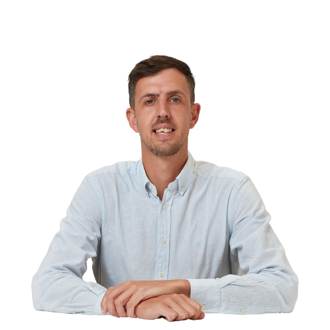A man sits with hands clasped on a table, wearing a light blue button-up shirt, against a plain white background.