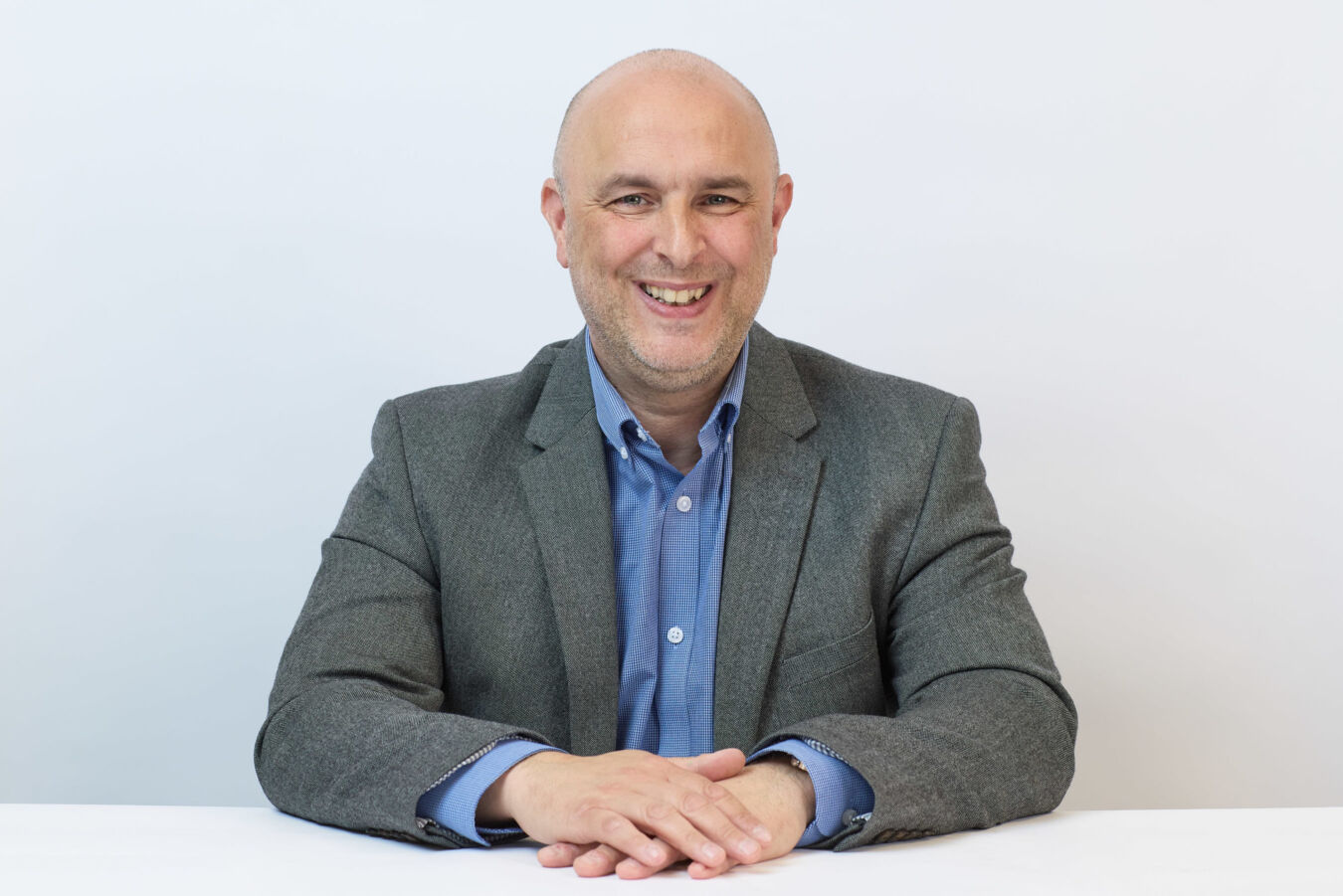 A person is smiling while sitting at a white table, wearing a gray blazer and blue shirt in front of a plain white background.
