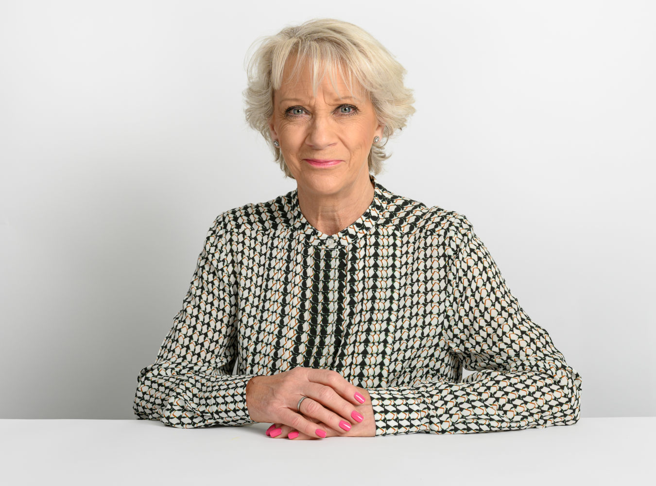 A woman sits with her hands clasped on a white table, wearing a patterned blouse and displaying pink-painted nails, against a plain white background.