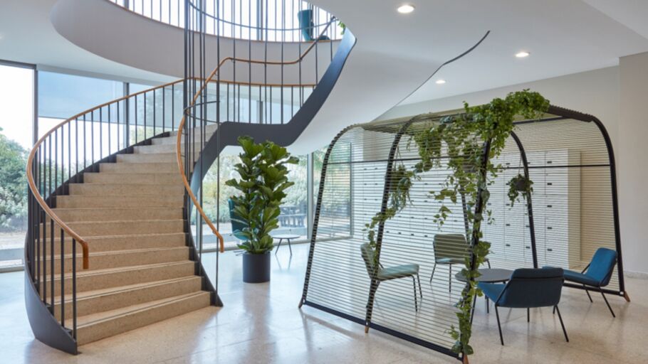Modern office entrance with a curved staircase, indoor greenery, and a glass meeting pod with hanging plants."