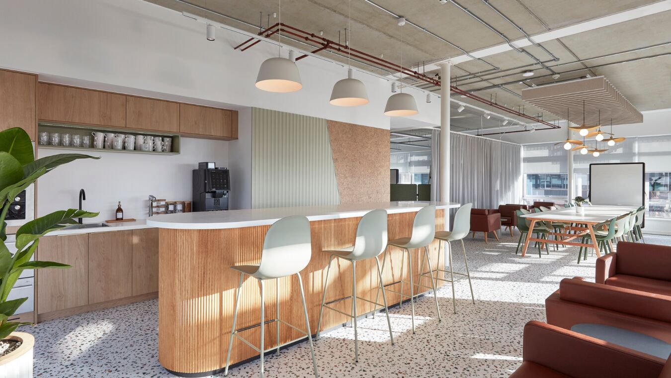 A modern office kitchen features a wooden counter with light green barstools and hanging lamps, surrounded by tables, chairs, sofas, and a whiteboard in a bright, open space.