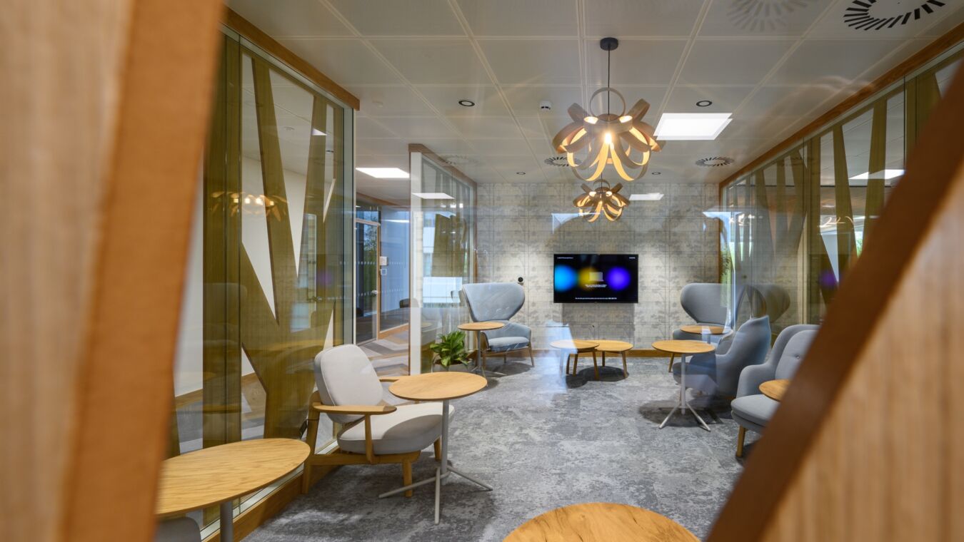 Modern meeting room with gray armchairs and round wooden tables, featuring abstract partitions and a glowing chandelier. A wall-mounted screen displays vivid colors