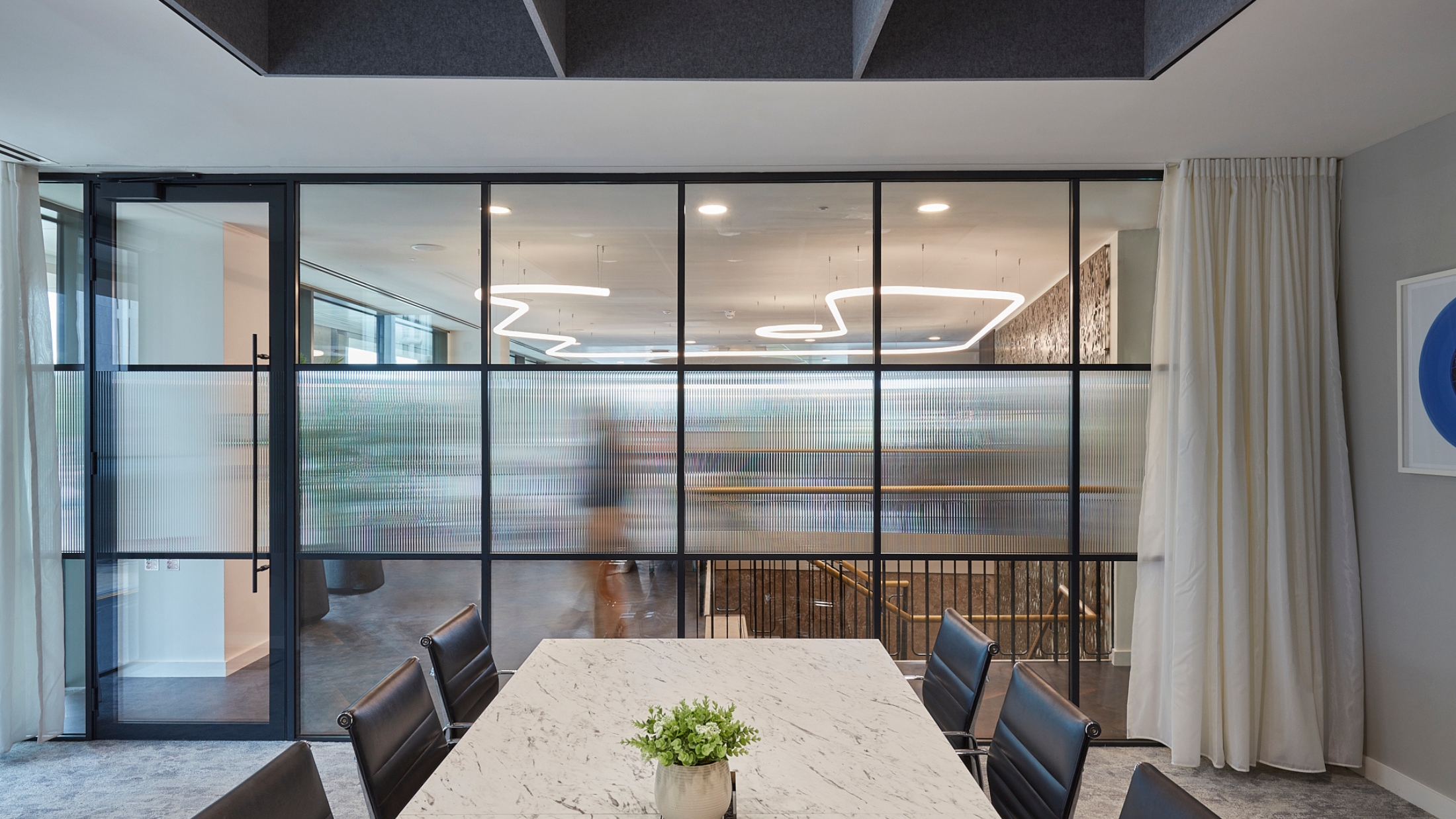 Conference room with a marble table and black chairs, facing frosted glass partitions. A blurred figure walks beyond the glass. Ceiling lights and wall art create a modern ambiance.