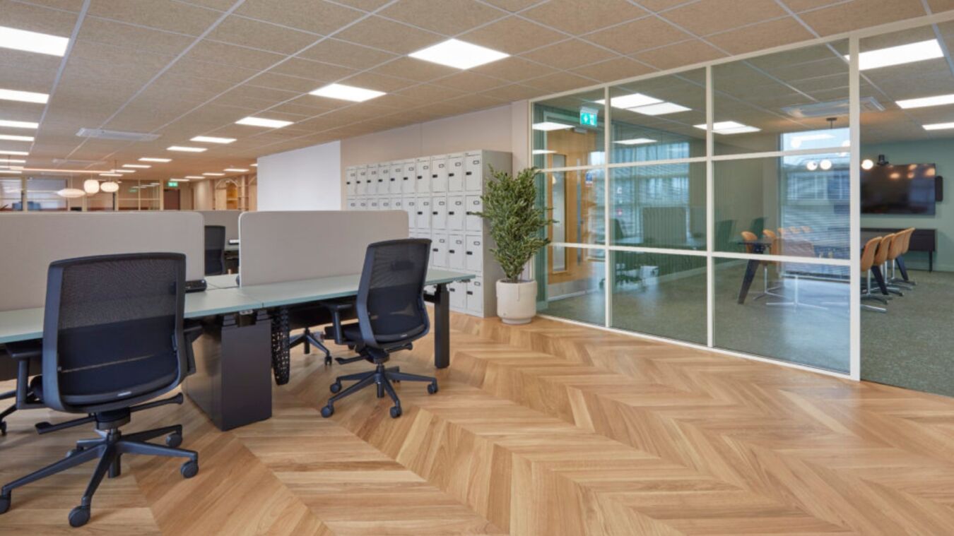 Office chairs surround desks with partition screens, placed on herringbone-pattern wood flooring; glass wall separates meeting room with chairs and screen, plant decorates workspace.
