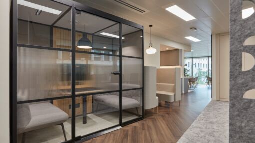 Modern office interior featuring a glass-enclosed meeting pod with bench seating and a pendant light, alongside open lounge seating and wooden flooring