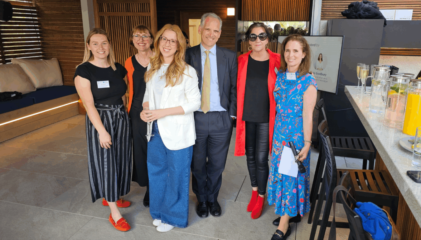 Six people stand smiling in a social gathering setting with wooden panel walls, next to a counter with glasses and drinks. A screen displays the text "Kylie Bradbury, Media Manager."