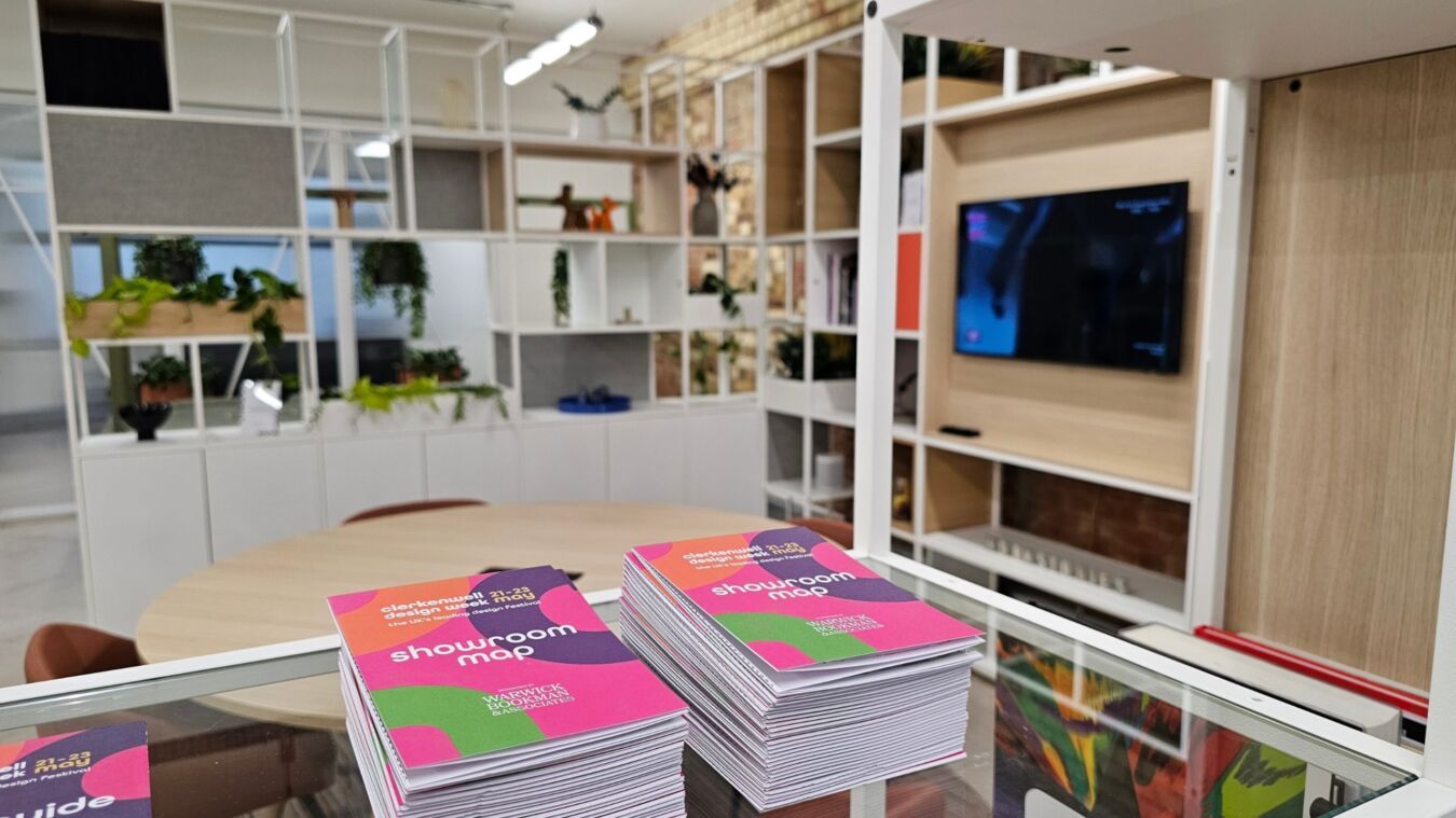 Stacks of "showroom map" brochures rest on a glass counter in a modern showroom, featuring white shelves with plants and a wall-mounted TV.