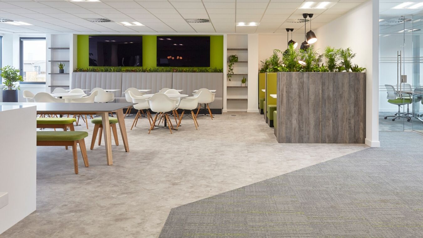 Modern office lounge with white chairs, wooden tables, and green accents. Plants divide spaces, creating a fresh and inviting atmosphere.