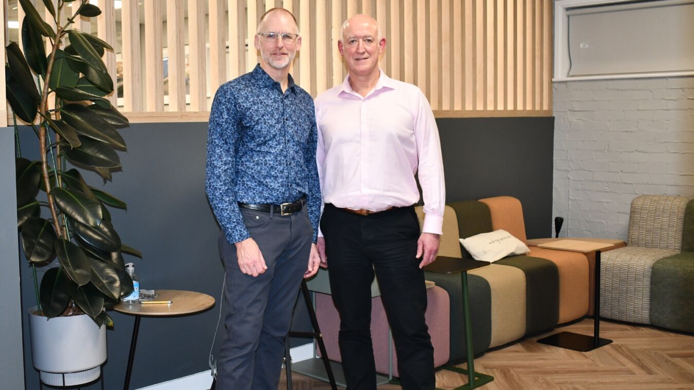 Two men stand smiling in an office with modern décor, featuring a large plant, wooden slat wall, colorful seating, and small tables.