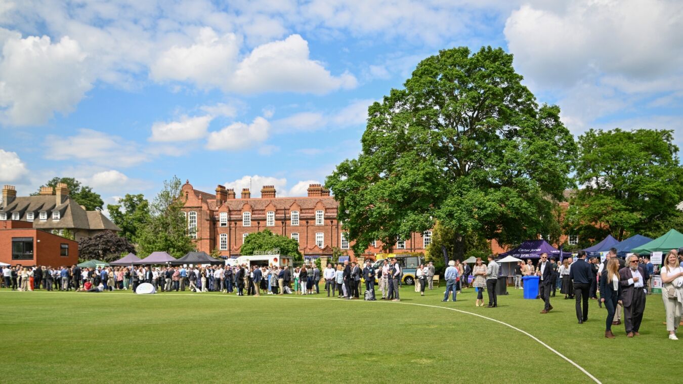 Crowd of people mingling during an outdoor event on a grassy field with large trees and historic red-brick buildings in the background, under a partly cloudy sky.