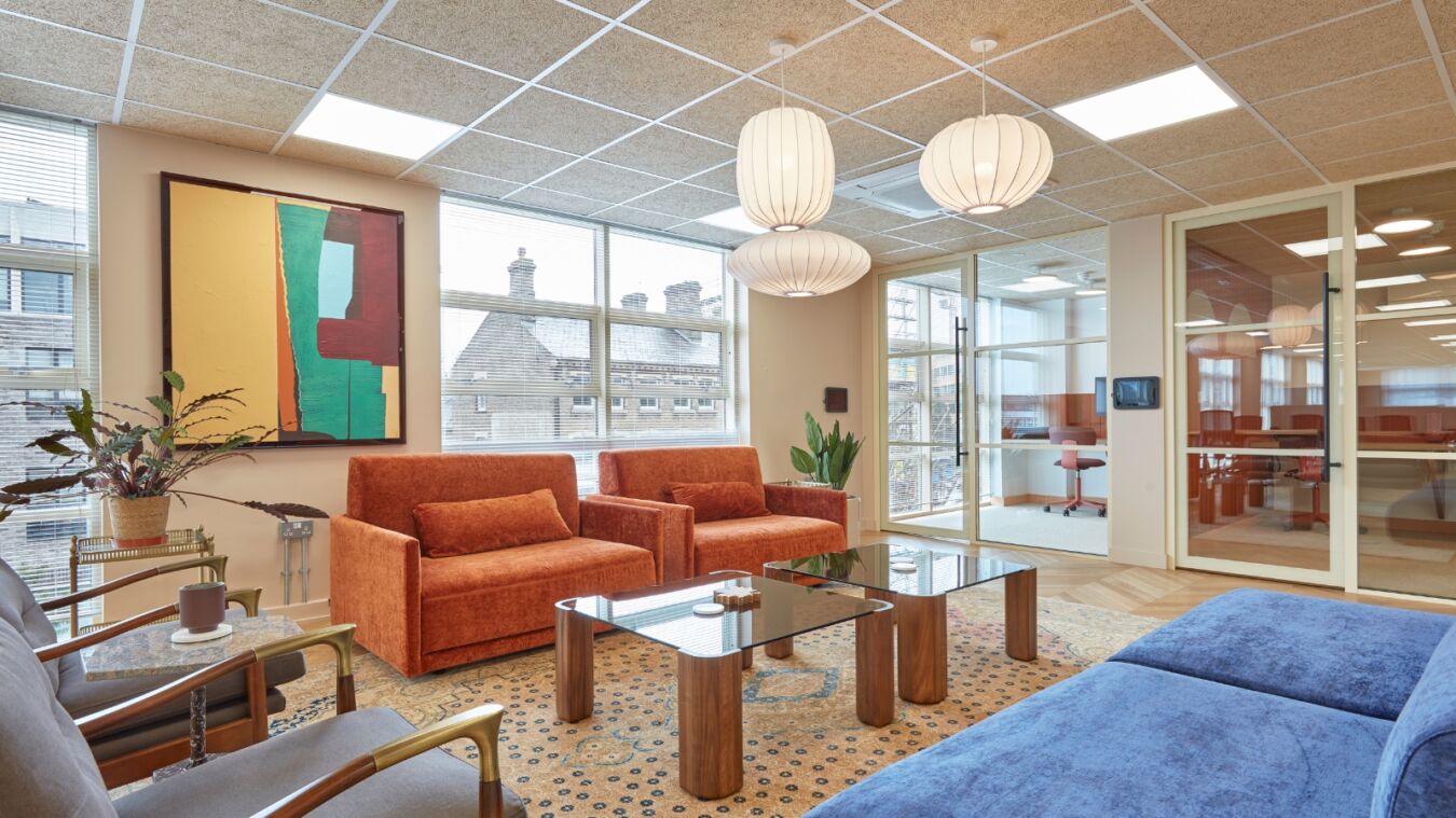 Stylish office lounge with orange armchairs, glass coffee tables, pendant lights, and large windows overlooking rooftops.