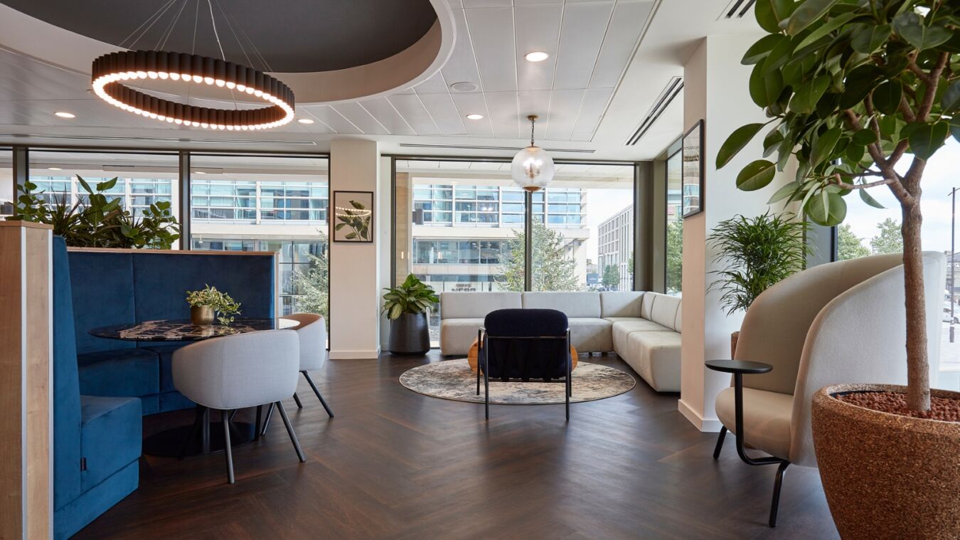 Modern office lounge featuring a circular chandelier above elegant seating, with plants and contemporary furniture near large windows showcasing an urban landscape.