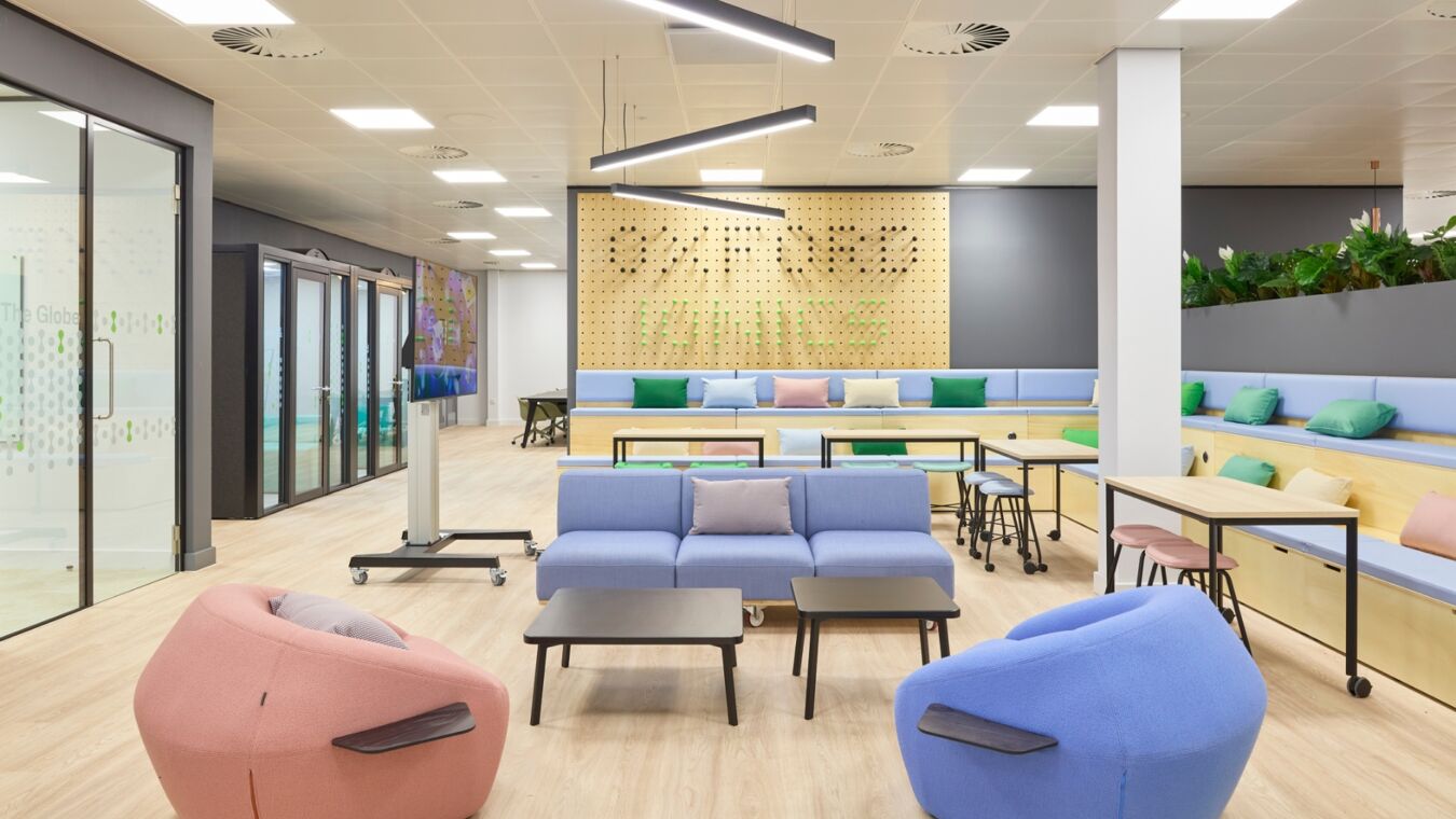Modern office space features colorful seating and small tables, with a wooden wall displaying "STEP INSIDE" in greenery; glass meeting rooms and a plant-lined wall surround the area.