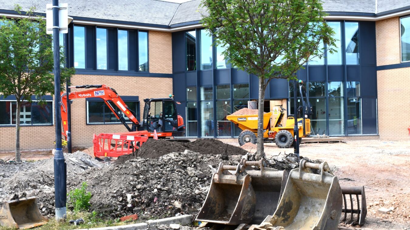 Construction machinery, including a Kubota excavator and a Thwaites dumper, operates on a site with dirt piles and a tree, in front of a modern brick building.