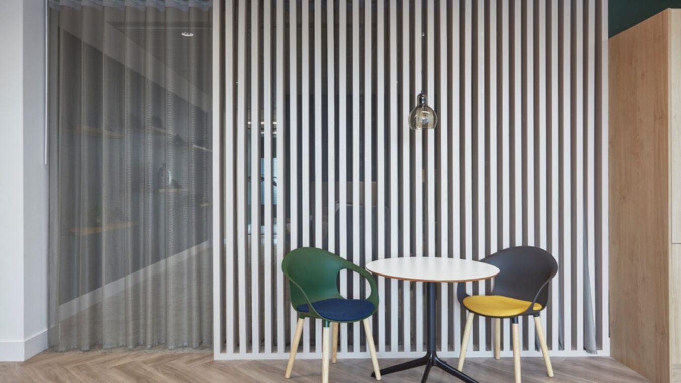 Two chairs, one green and one gray, face a round white table in front of vertical wooden slats, with a hanging light bulb above, in a modern room.