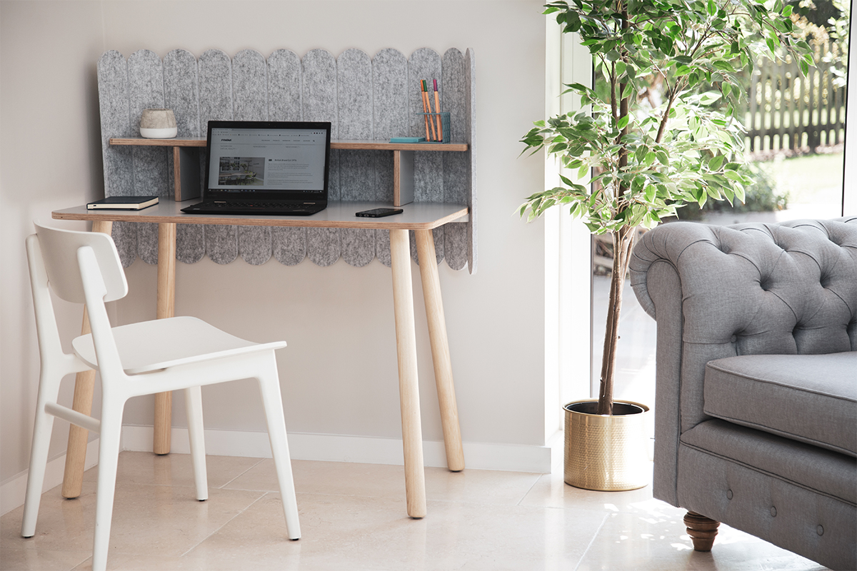 Wooden desk with a laptop displaying a website, set against a gray felt backdrop, accompanied by a white chair. Nearby, a potted plant and gray sofa enhance the bright room.