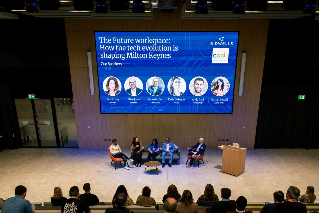 Panel discussion takes place in an auditorium, with speakers seated on stage. A large screen overhead reads, “The Future workspace: How the tech evolution is shaping Milton Keynes.” Audience listens attentively.