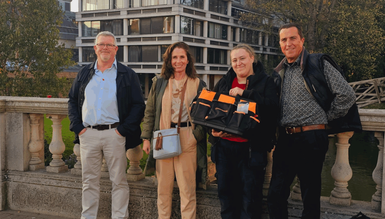Four people stand on a stone bridge; one holds a black and orange tool bag. Behind them, a modern building and trees are visible.