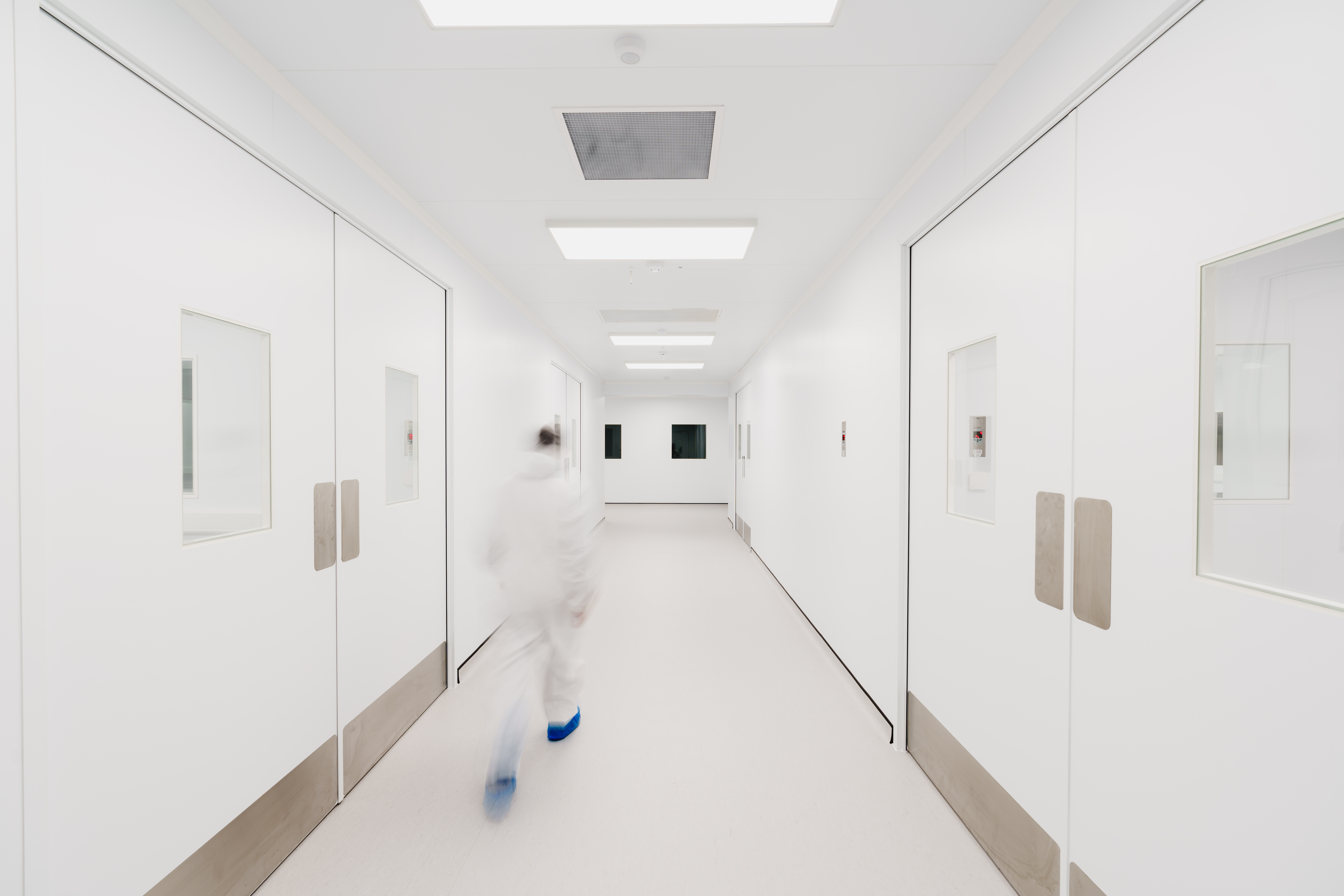 Person in protective clothing walking down a pristine, white corridor with large doors, bright lighting, and a sterile environment, suggesting a laboratory or cleanroom setting.