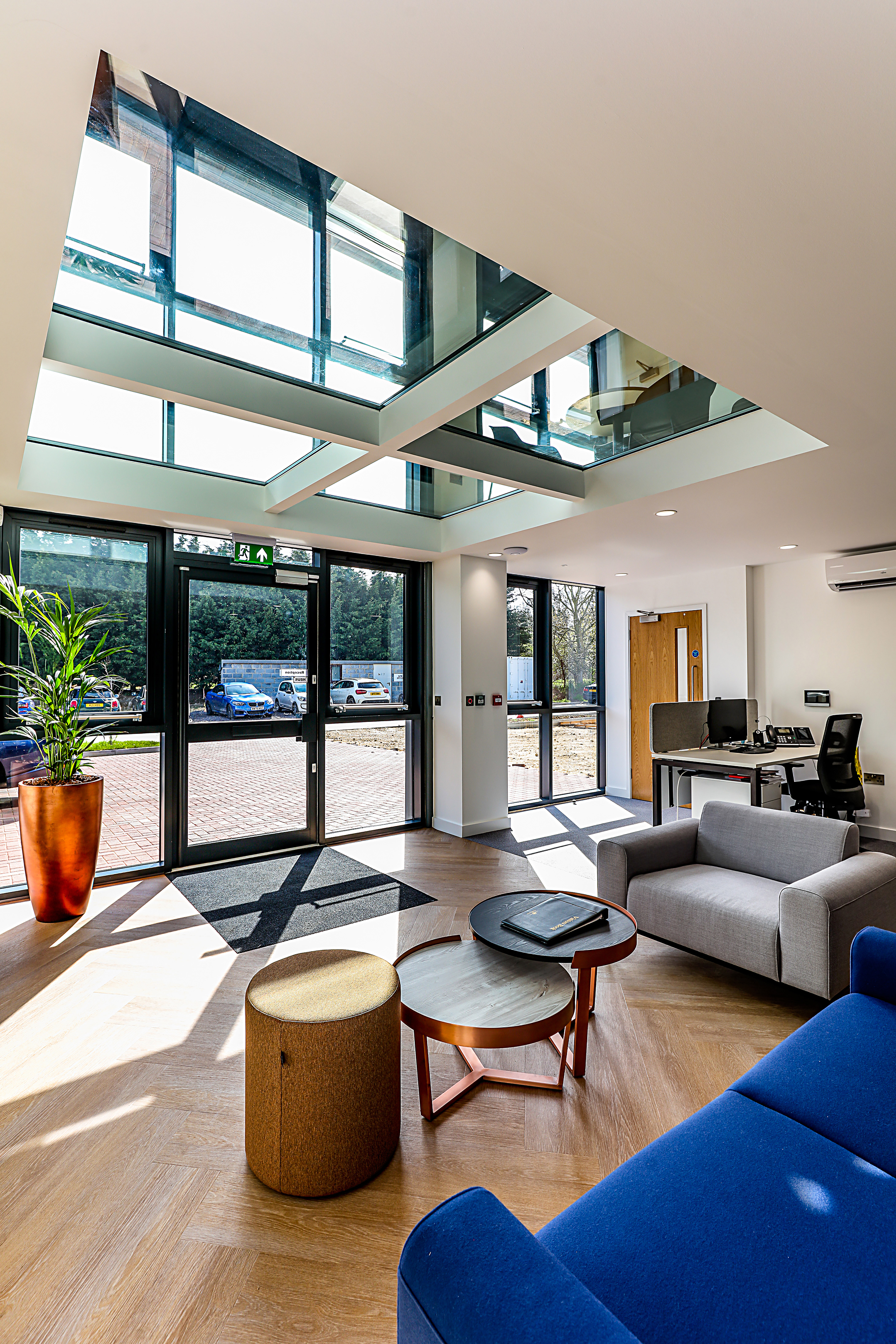Spacious office lobby features blue and gray seating, a round ottoman, nested tables, and a potted plant; large glass doors and ceiling windows let in natural light, overlooking a parking area.