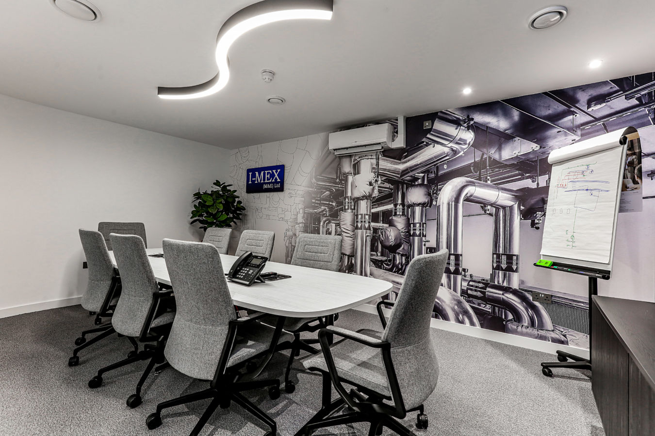 Modern conference room with a white meeting table, grey office chairs, a wall-mounted screen, and a feature wall displaying a large industrial piping graphic