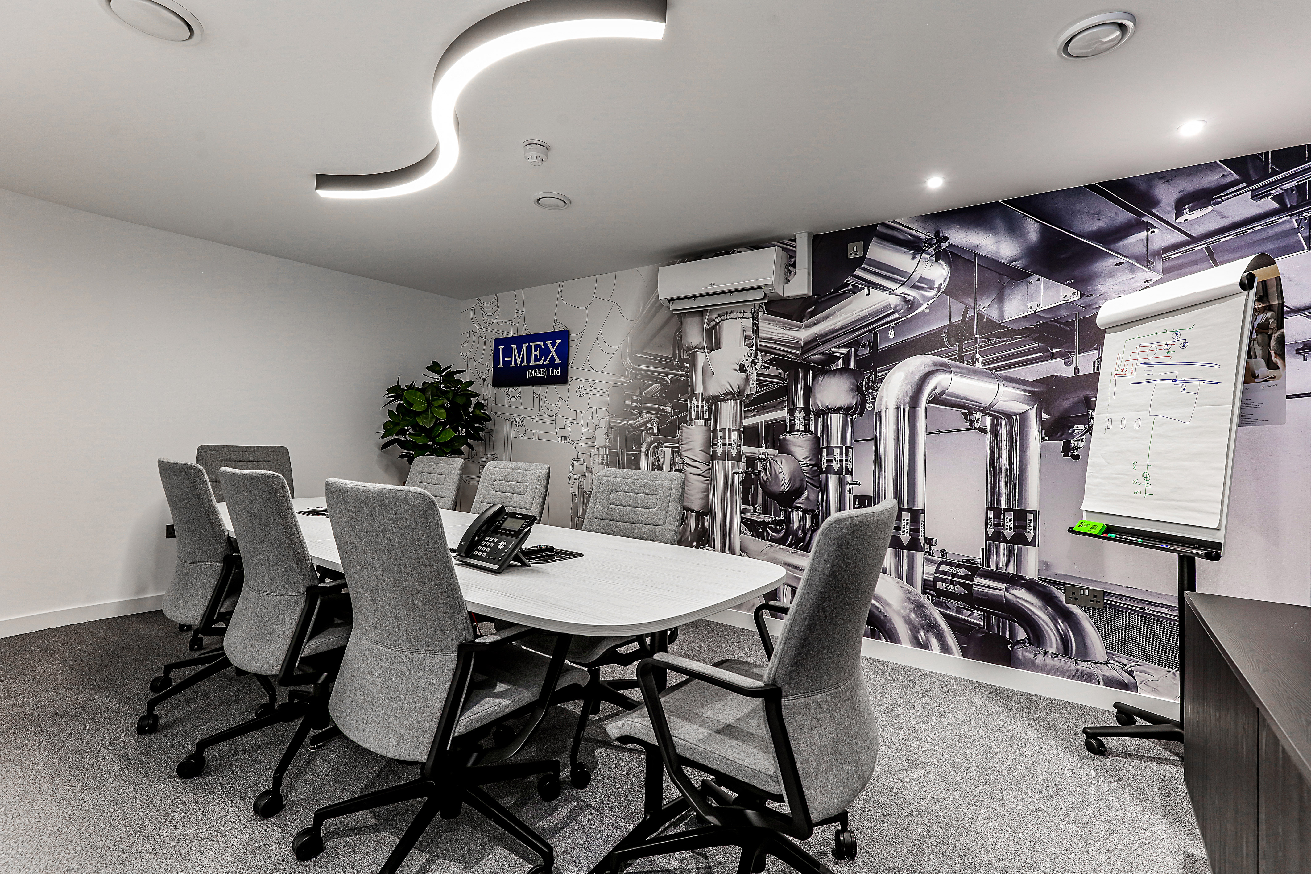 Modern conference room with a white meeting table, grey office chairs, a wall-mounted screen, and a feature wall displaying a large industrial piping graphic