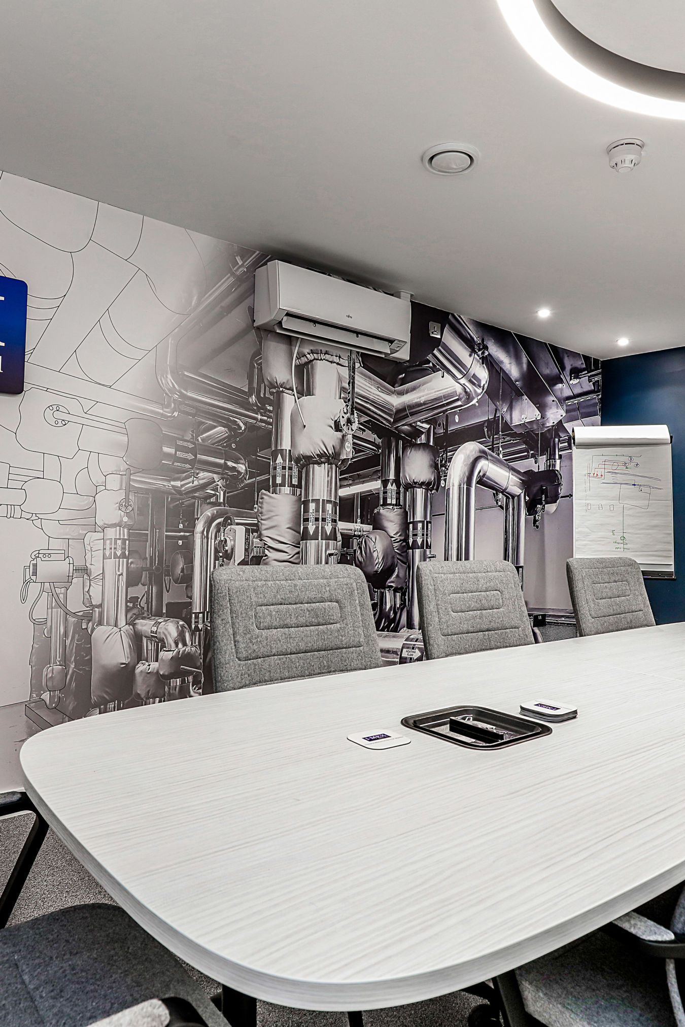 Conference room with a large oval table surrounded by gray chairs, against a wall mural of industrial pipes and machines; a flip chart with diagrams stands nearby, creating a modern, professional atmosphere.