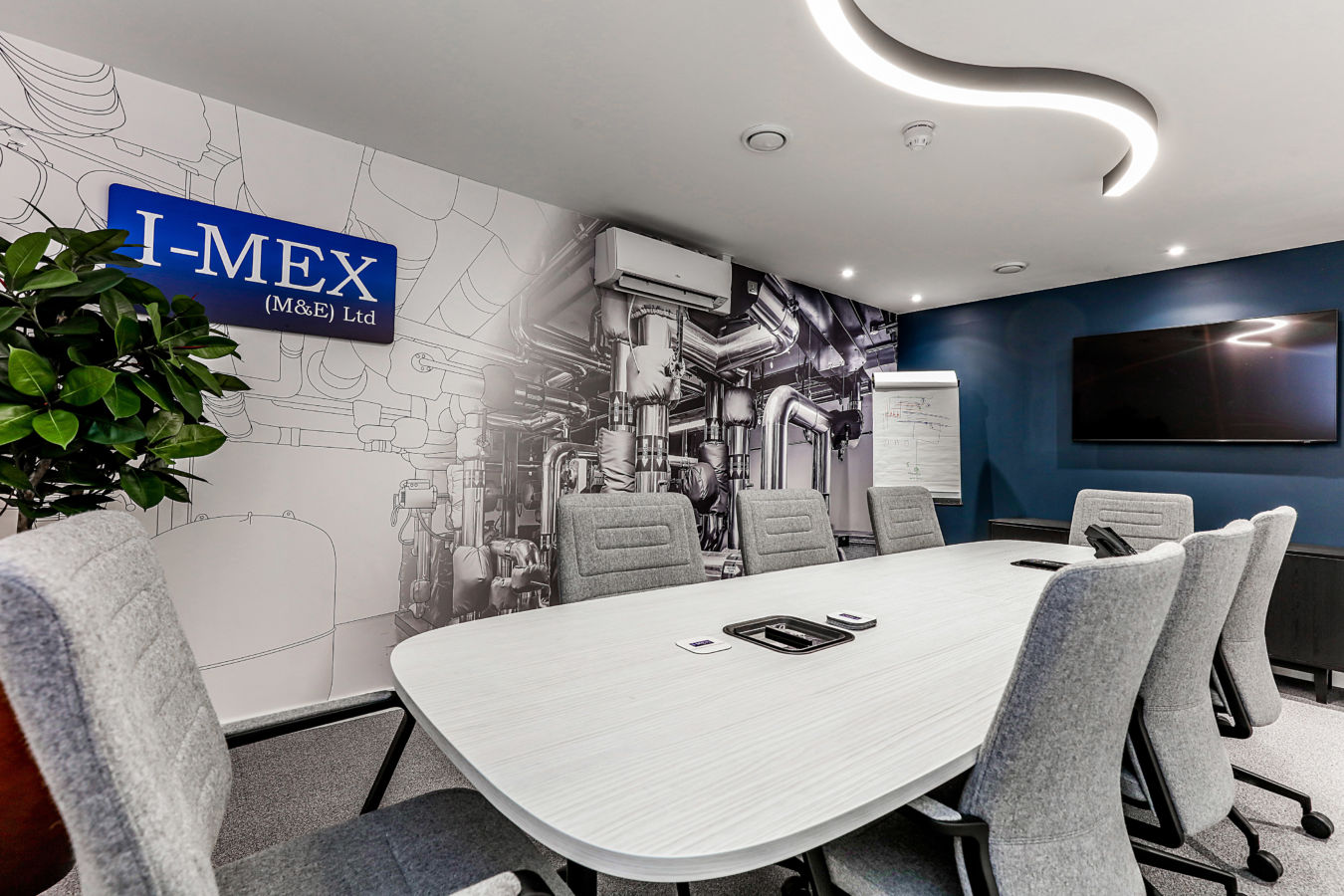 Conference room with a large oval table surrounded by gray chairs, against a wall mural of industrial pipes and machines; a flip chart with diagrams stands nearby, creating a modern, professional atmosphere.